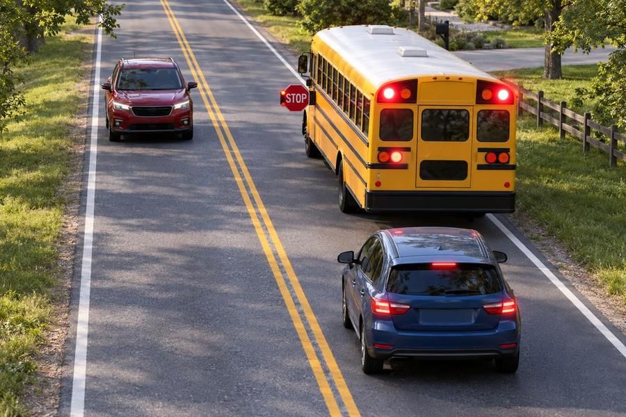 A school bus with flashing red lights and its stop sign out on a two-lane road, with a car stopped behind it and another car stopped in the opposite lane